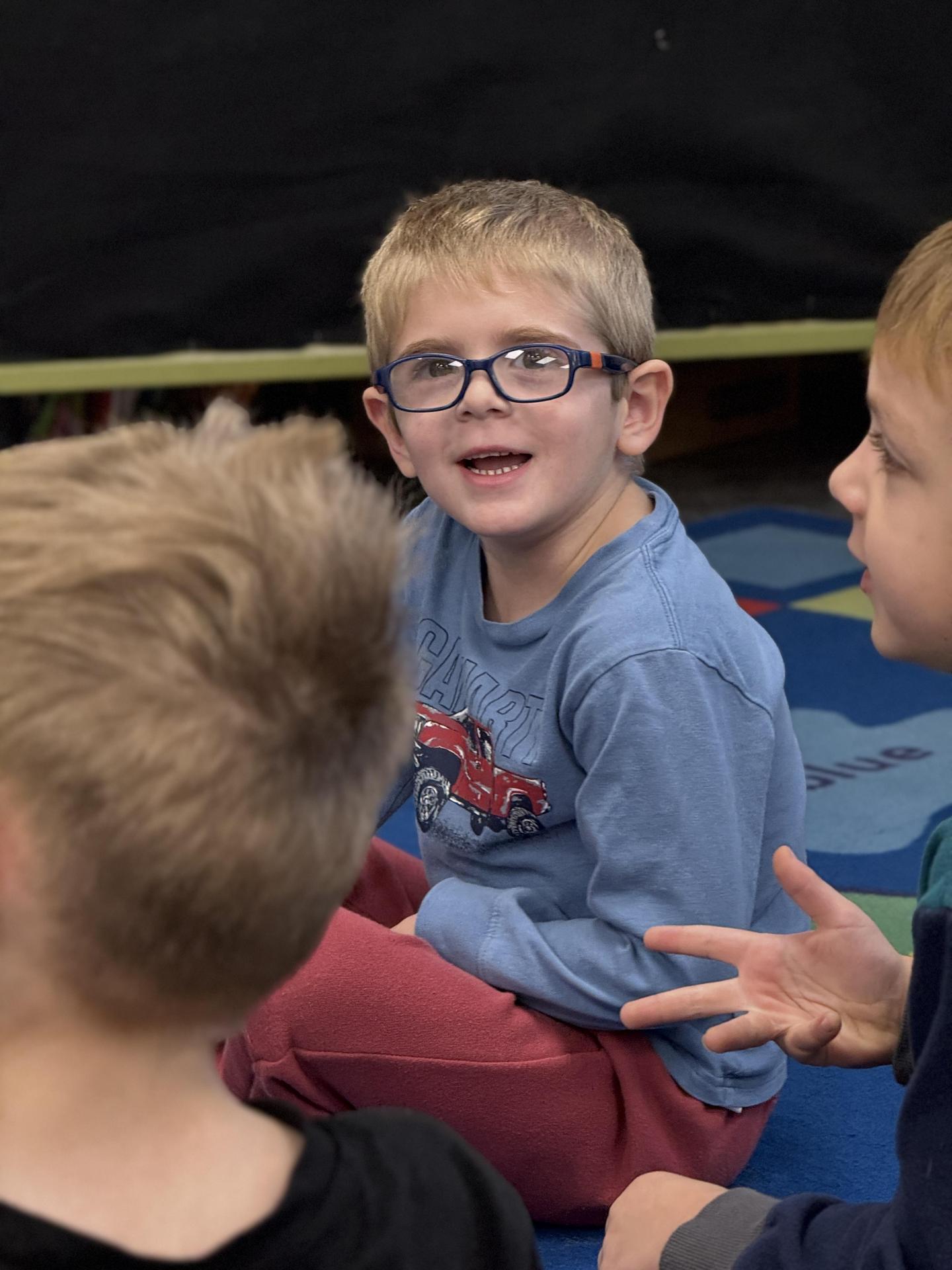 Young boy with glasses smiling and sitting on a colored rug with peers.
