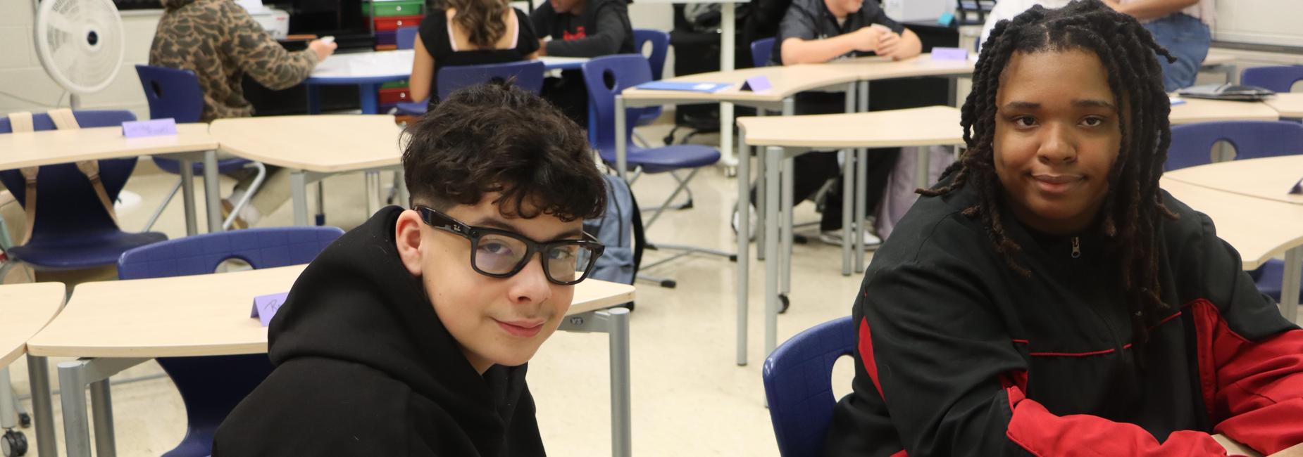 Two students sitting at a classroom table, smiling at the camera.
