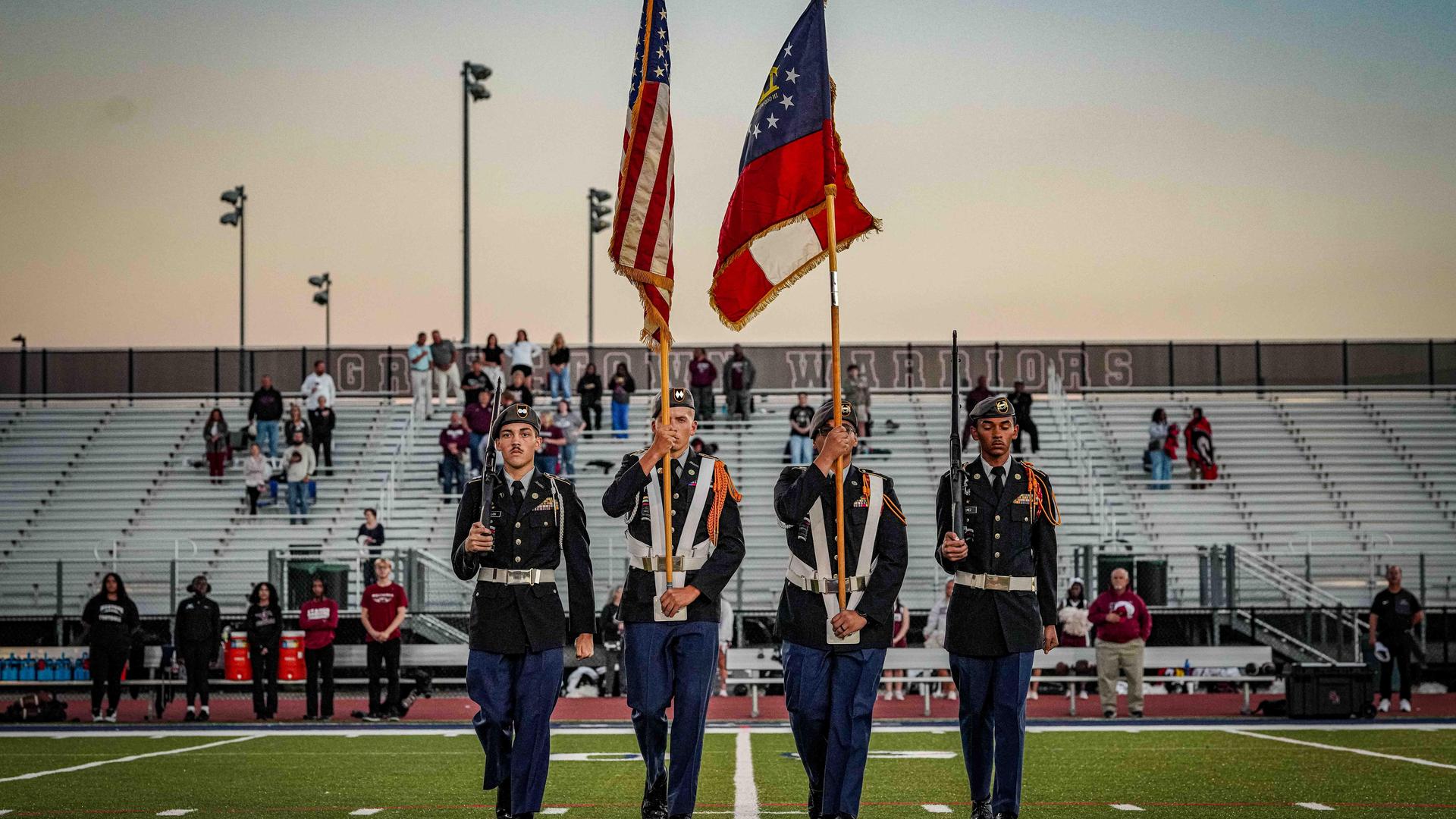 Four people in military dress uniforms, holding flags, marching across a football field.
