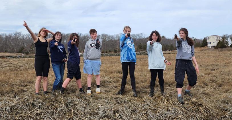 Seven young people posing and pointing in a field against a cloudy sky.