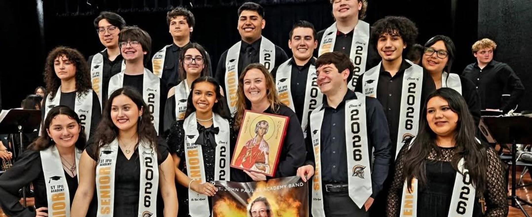 Group of students in formal attire posing with a banner and a portrait.