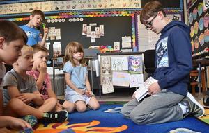 Boy reading a book to a group of attentive children on a colorful rug.