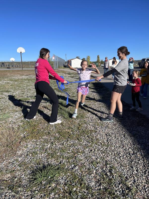 Young girl running through a finish line with support from two trainers during an outdoor event.