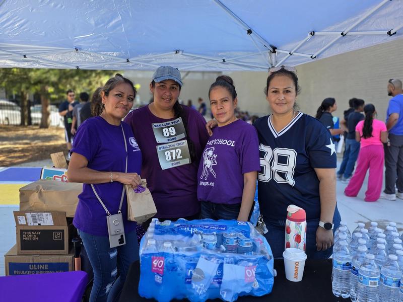 Four women in purple shirts gather under a tent with drinks and snacks.