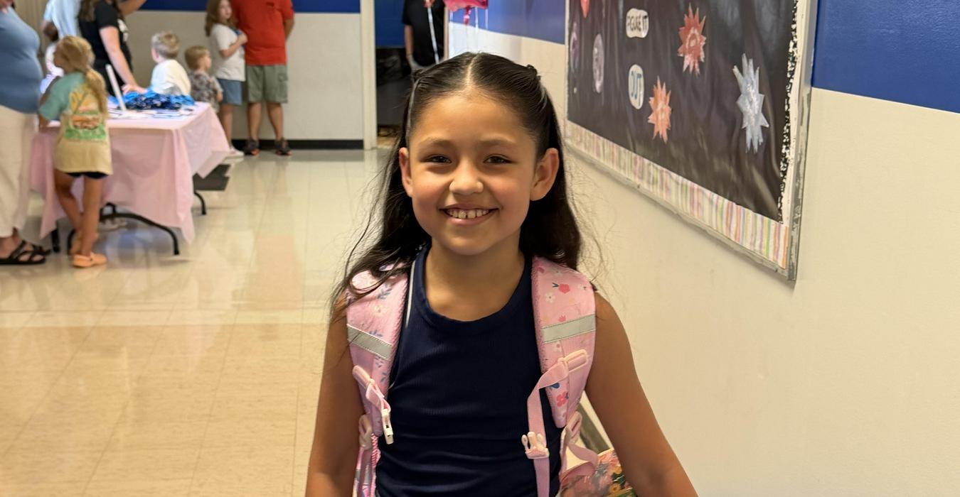 A girl holding a box of crayons while wearing a backpack in a hallway.