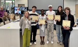 A group of adults and students stand together with certificates during a meeting