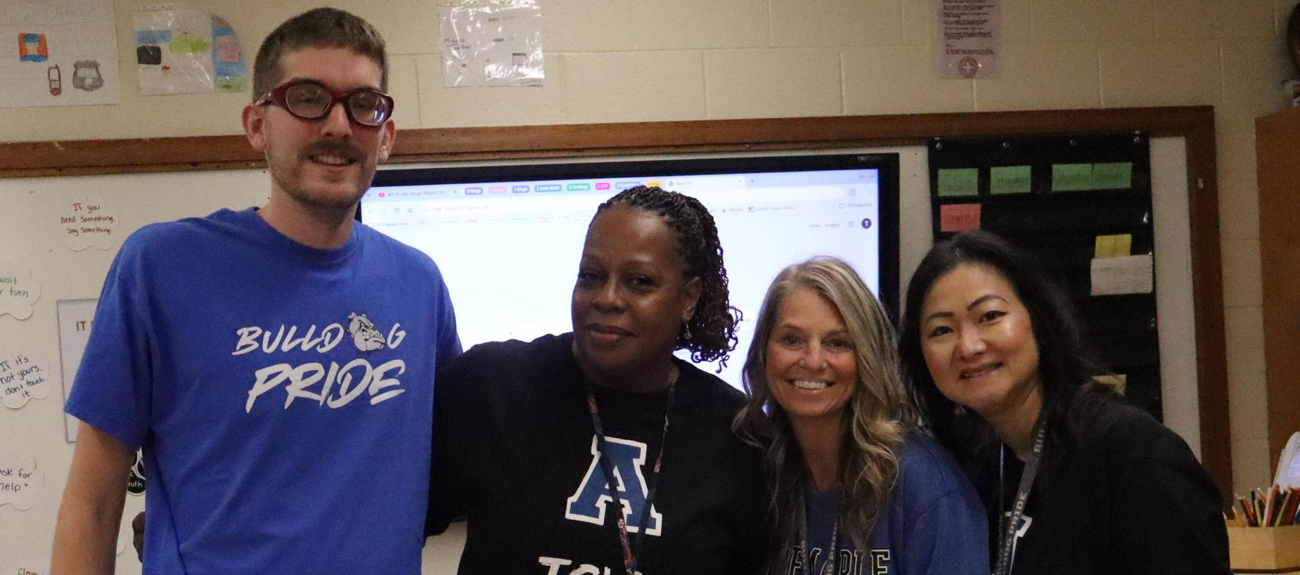Four people smiling in front of a screen, wearing blue and black team spirit apparel.