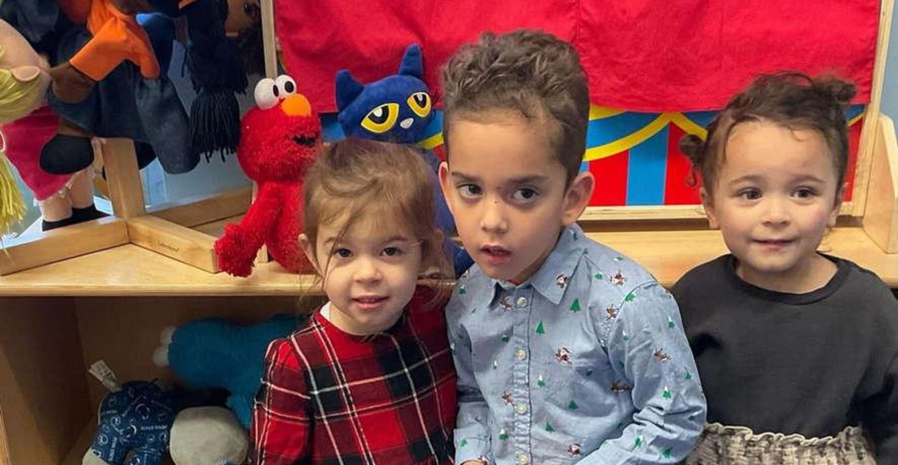 Three children posing in front of a wooden shelf with toys.