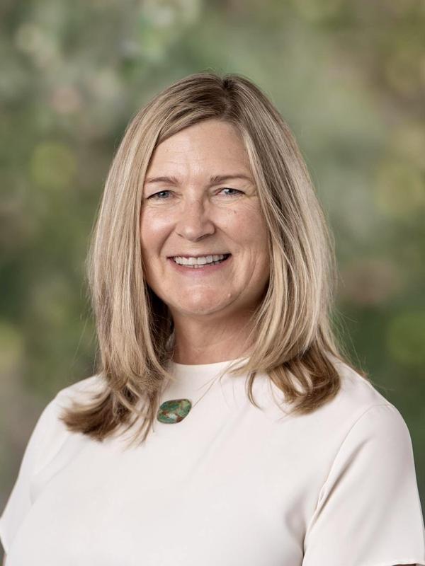 A smiling woman with long, straight hair and a green necklace against a blurred background.