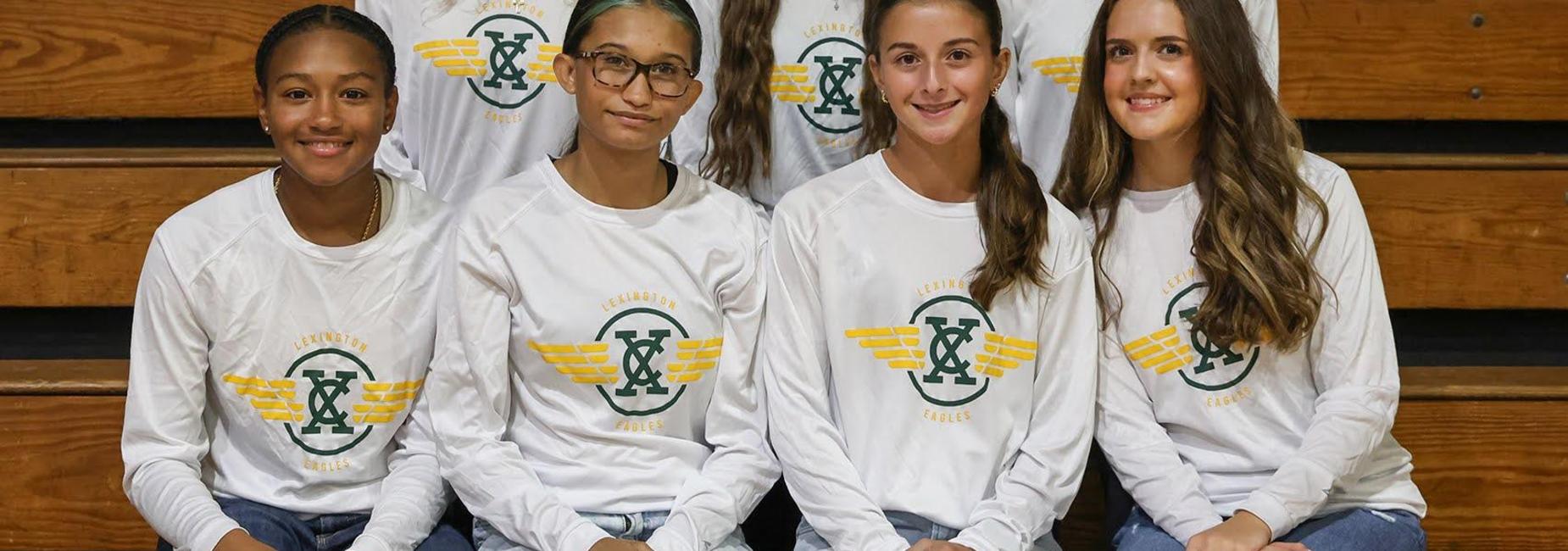 Group of young female athletes in white shirts sitting on gym bleachers.