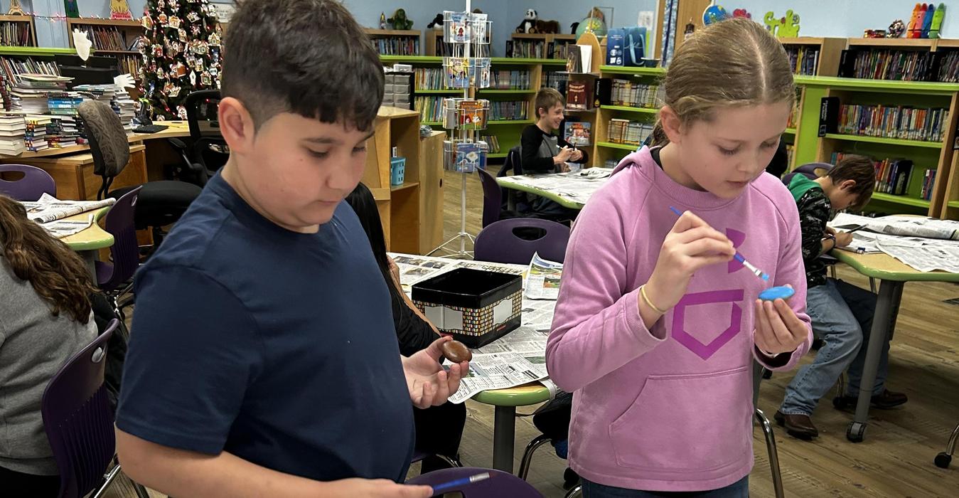 A child paints a small object while another child holds a paintbrush, surrounded by newspaper.