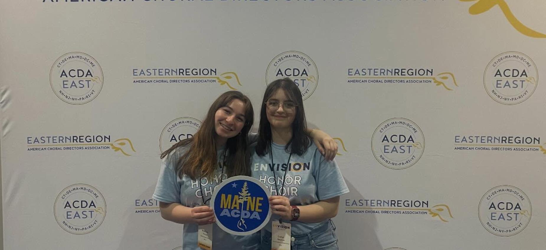 Two smiling girls holding a sign in front of a promotional backdrop.