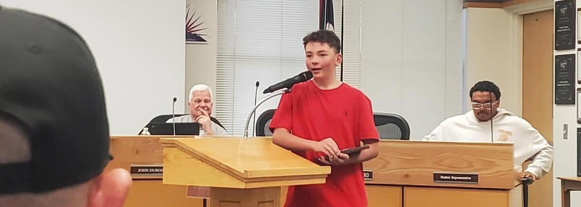 Teen boy speaking at a podium with two adults listening in a meeting room.