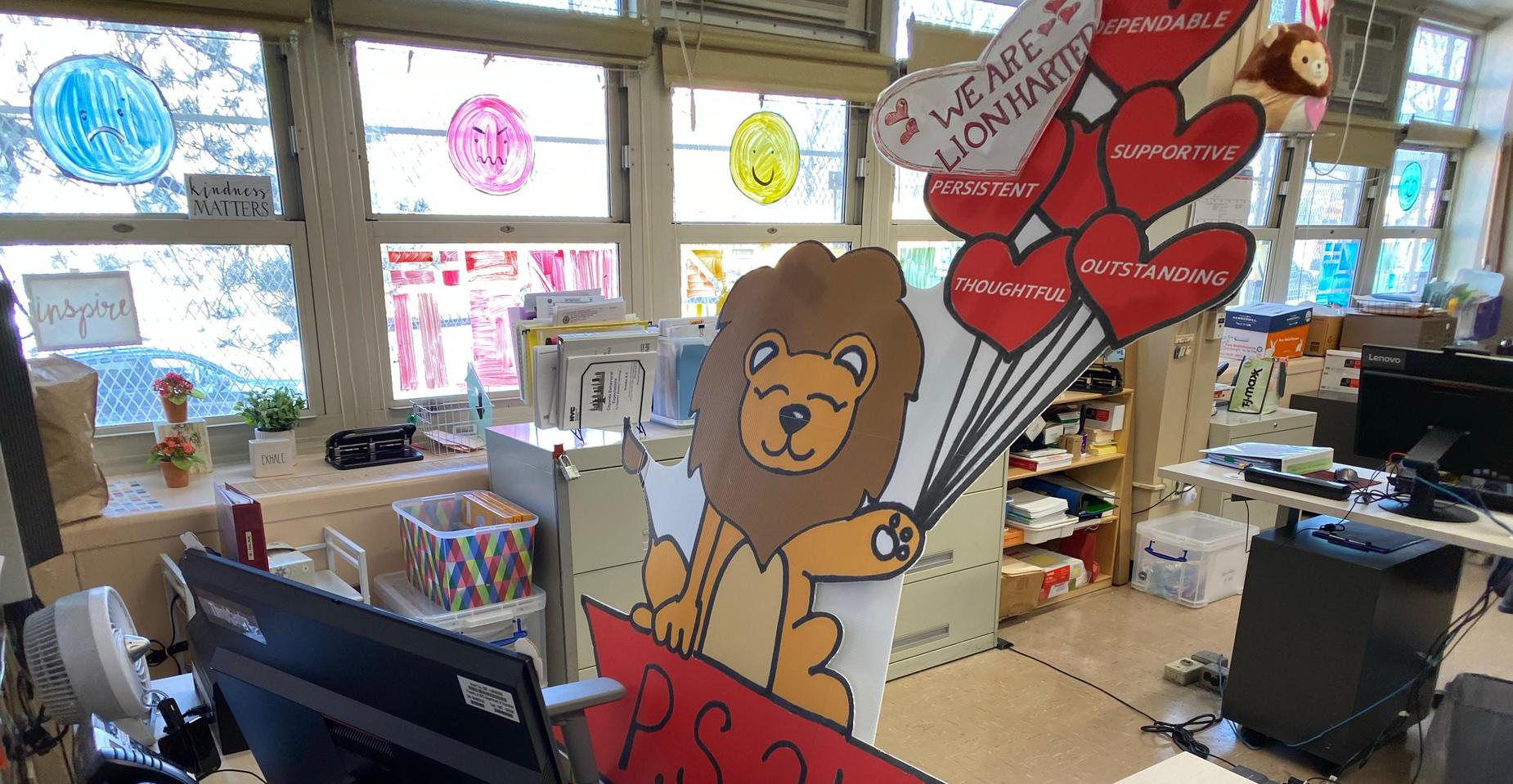 View of a lion mascot at a desk with a computer in a classroom setting.