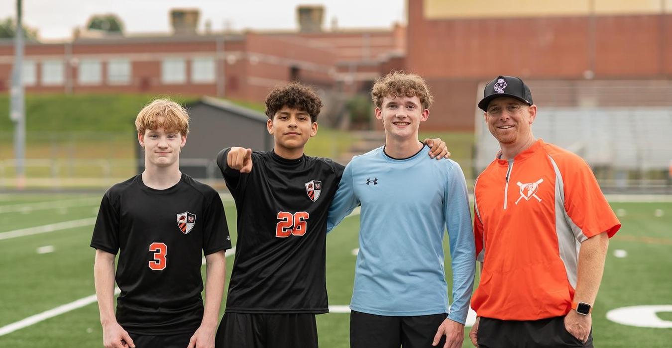 Four young male soccer players posing with a coach on a sports field.