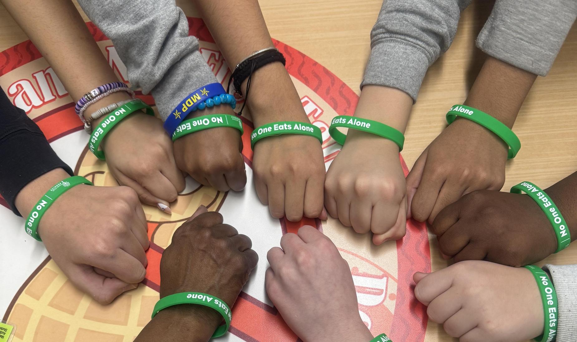 Several hands on a table with green bracelets with the words No One Eats Alone