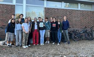A group of high school students holding a "Bike to School Week" flyer stand in front of a full bike rack