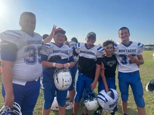 Five young football players in uniforms, smiling and posing together on a field.
