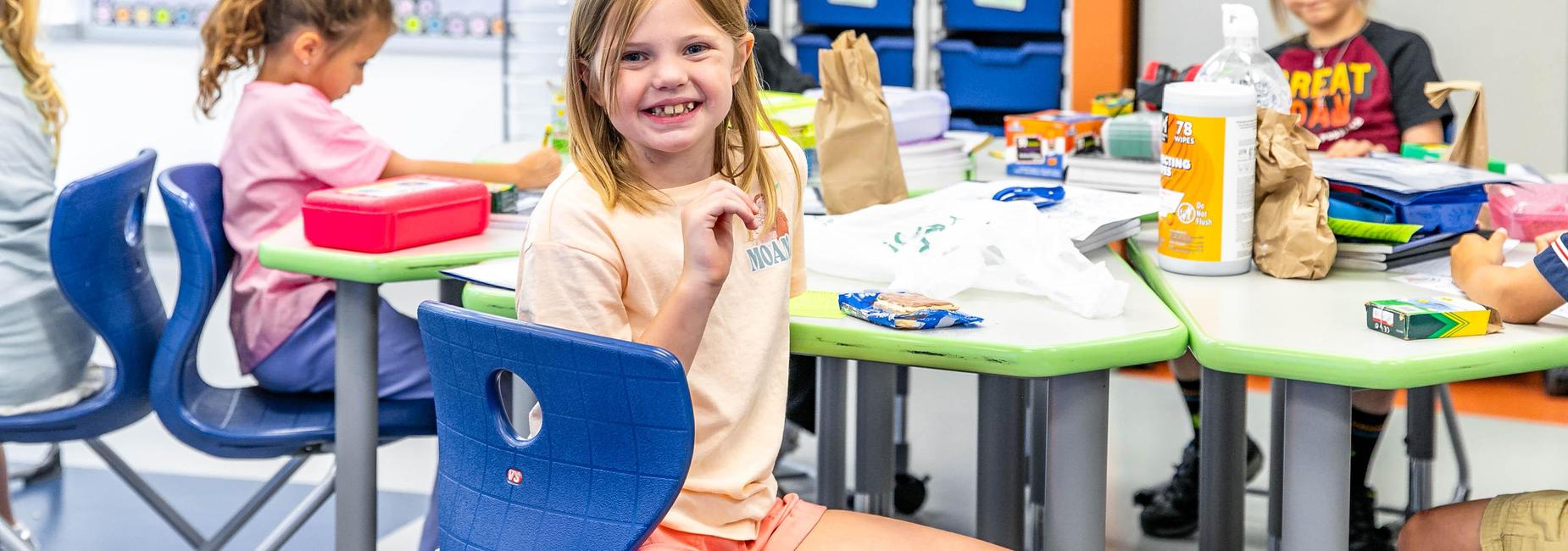 A girl smiling and enjoying snacks at a classroom table while classmates are busy.