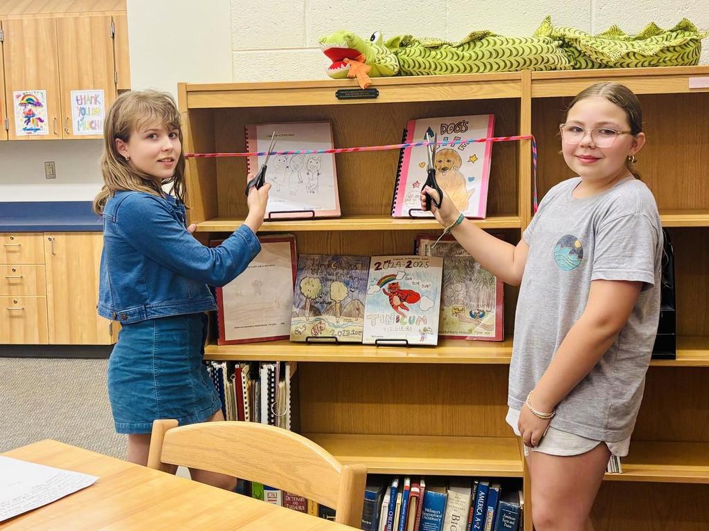 Two elementary students cutting a ribbon in the library for a new shelf displaying self-published books.