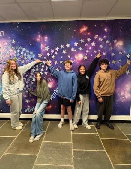 Five students pose in front of a starry mural, pointing towards a decorative display.
