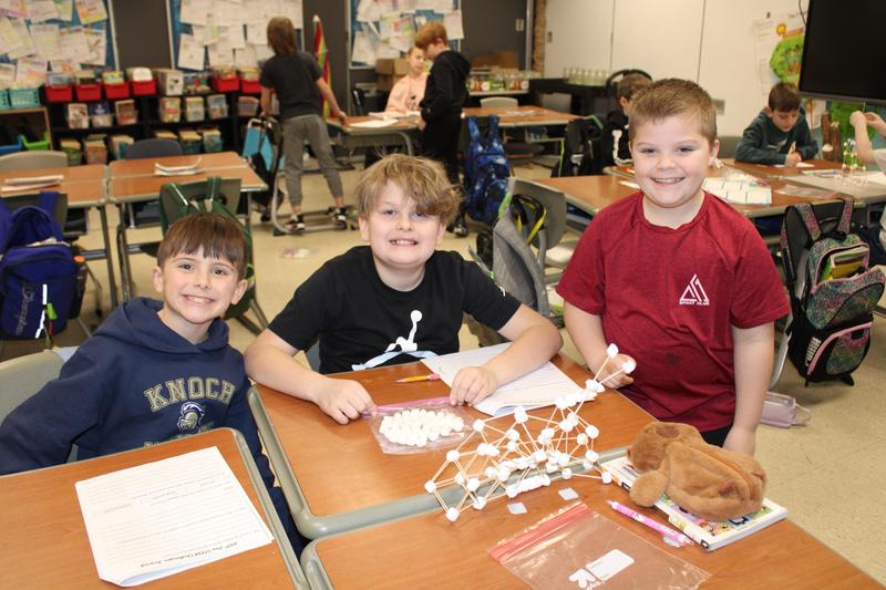 3 boys smiling with tower they built on desk in front of them