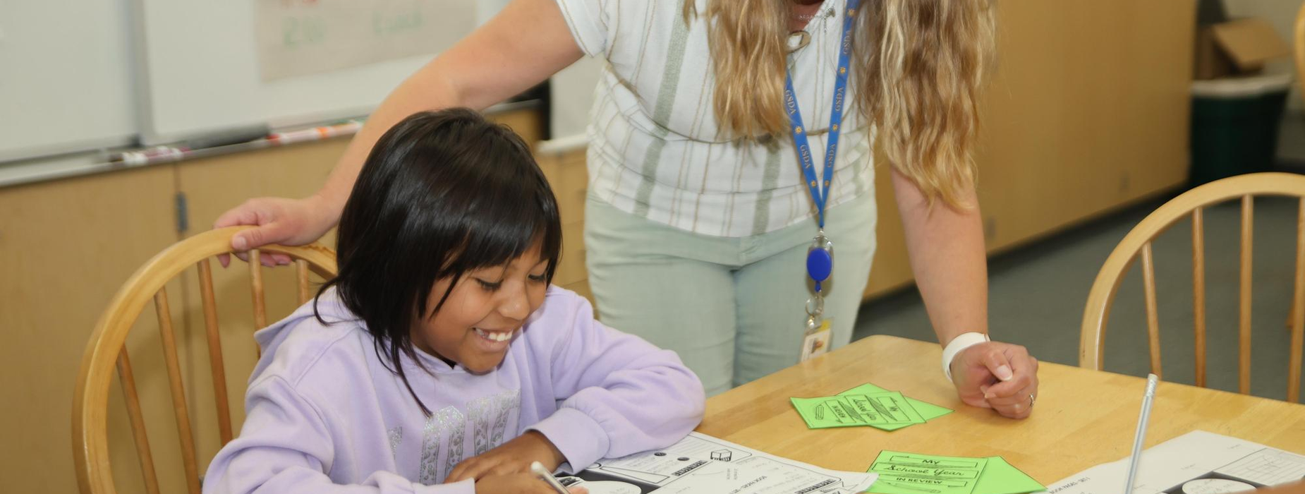 Teacher helping a smiling girl with schoolwork at a table.