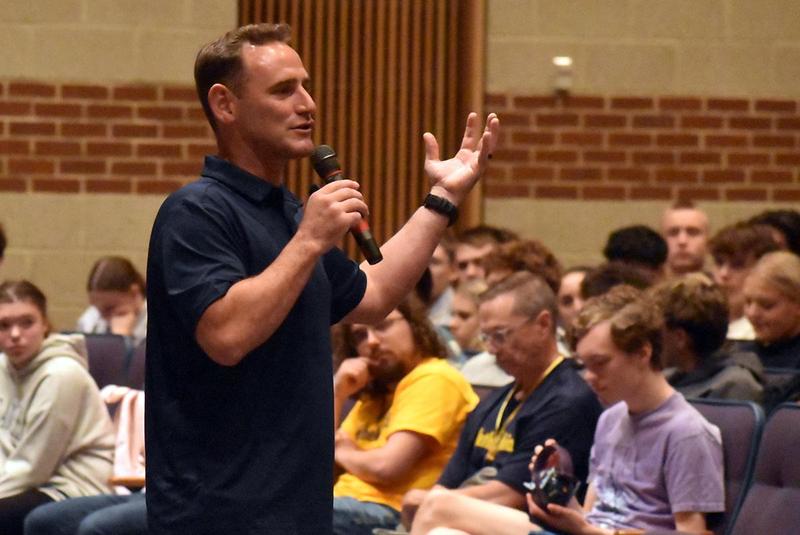 A speaker with a microphone gestures to an audience seated in a classroom setting.
