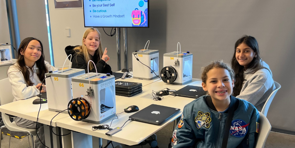 Four girls at a tech workshop, engaging with equipment and smiling at the camera.
