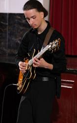 Boy with pulled back hair playing the bass, and boy playing the piano in the background
