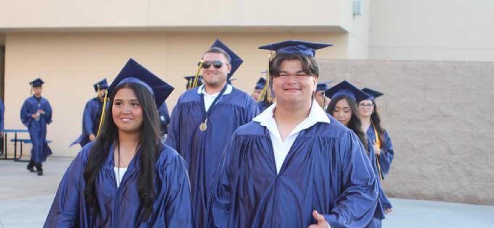 Students in blue gowns walking together, some holding drinks, on a school campus.
