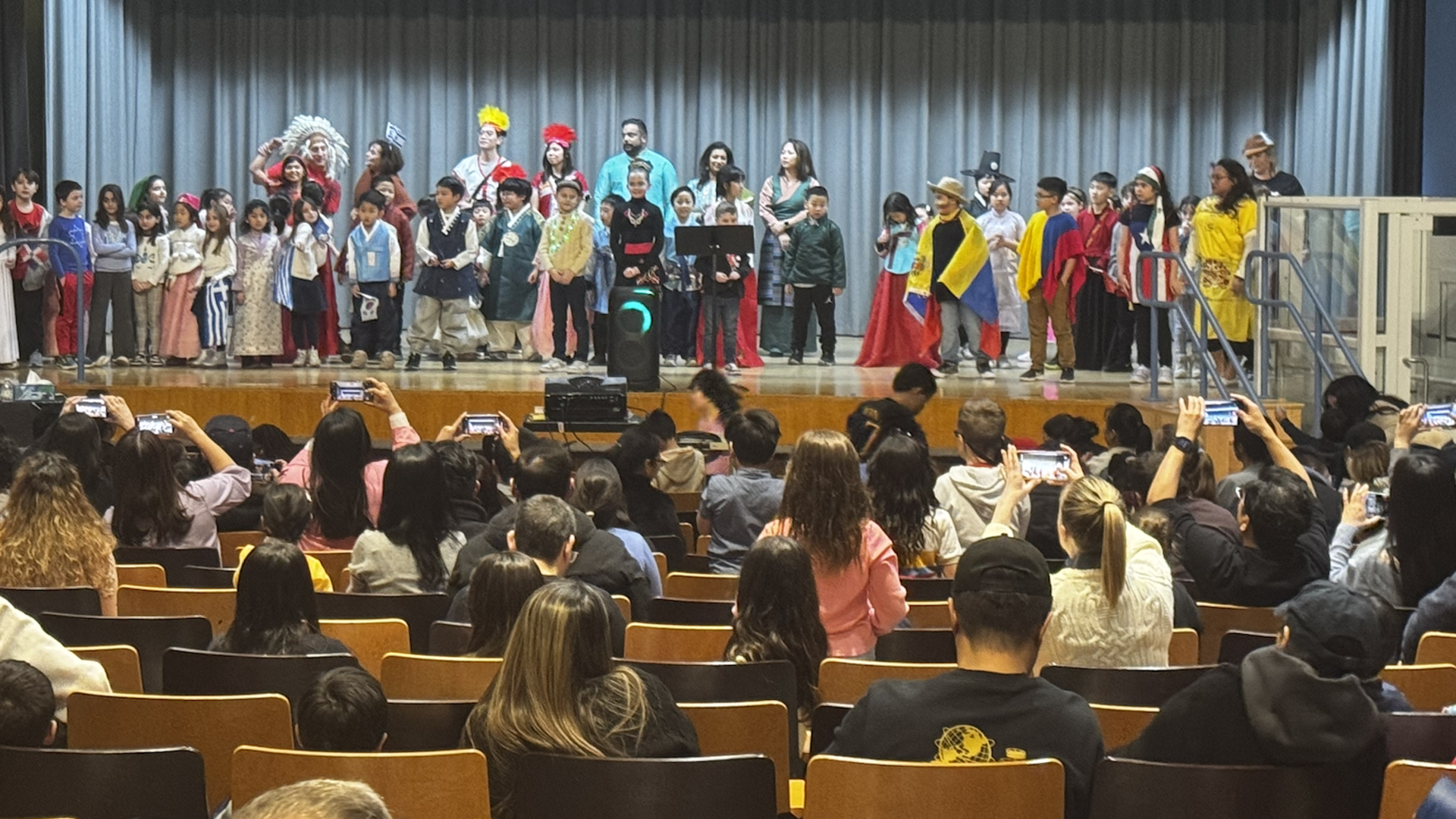 A diverse group of students in costumes perform on stage before an audience.