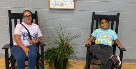 Two people sitting in rocking chairs near a potted plant and a family sign.