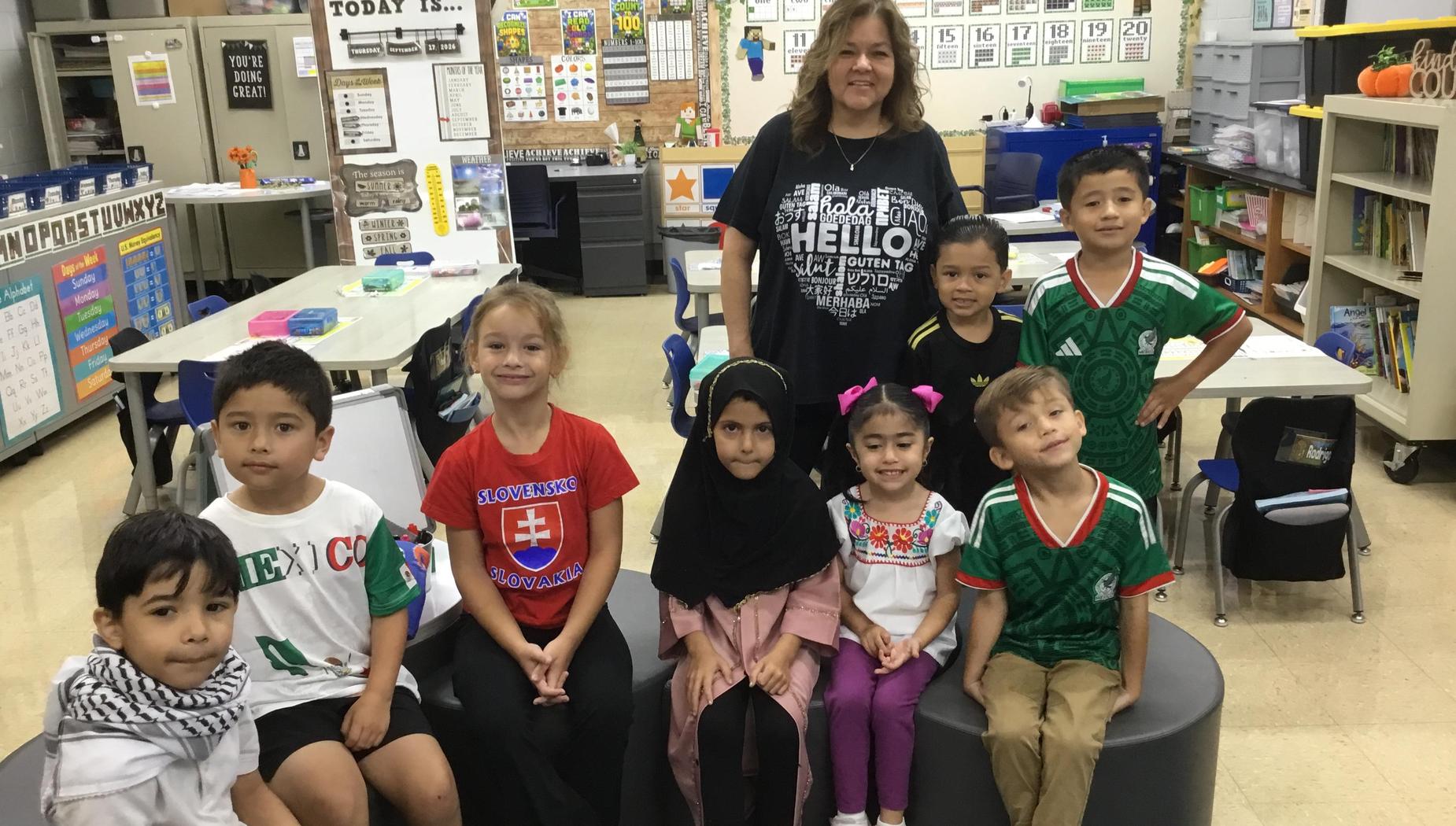 Eight children seated and standing in a classroom, including a teacher all in casual attire.