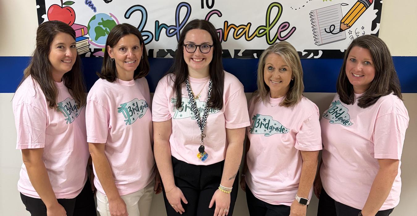 Five women in pink shirts stand in front of a colorful 'Welcome to 3rd Grade' sign.
