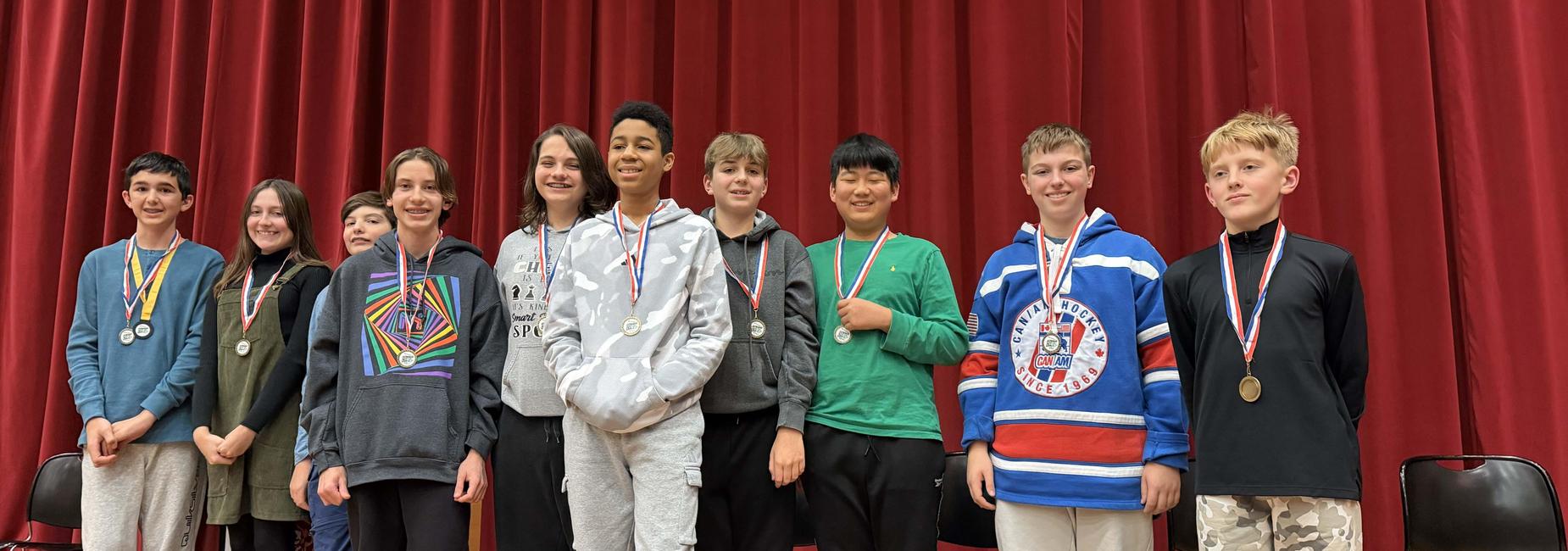 A group ten geography bee contestants standing together, wearing medals and smiling on stage.