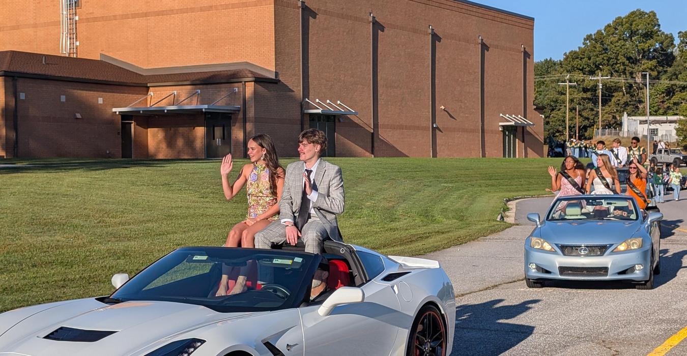 A couple waves from a convertible car during a parade, with others in the background.