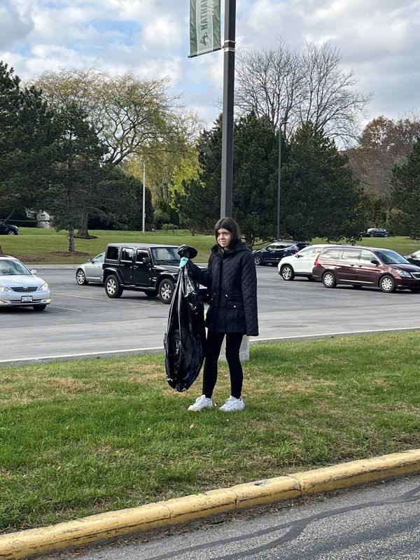 A girl standing on the grass holding a garbage bag in a parking lot.
