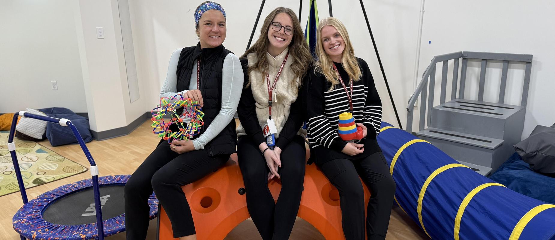 three occupational therapists smile in their OT room with their various gadgets for therapy