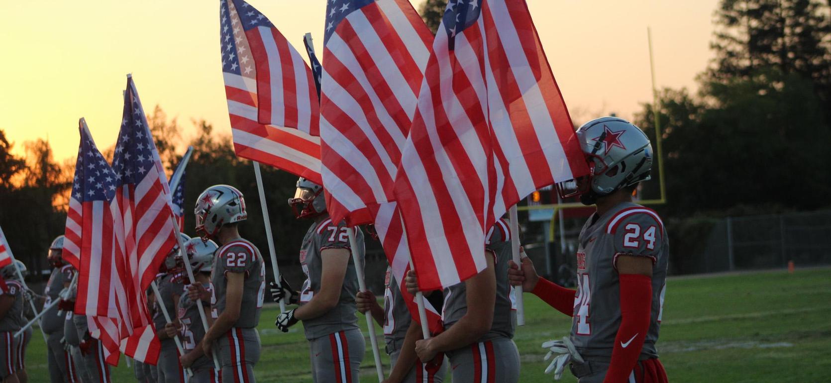 Football players in grey uniforms holding American flags on a field at sunset.