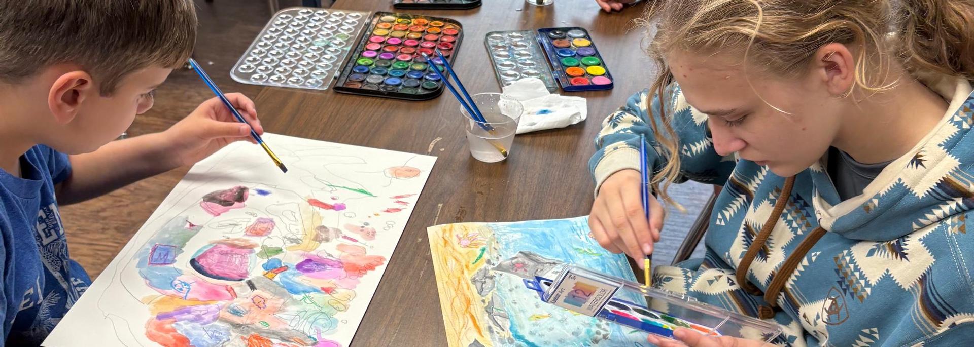 Two children creating art at a table, using watercolors and brushes.