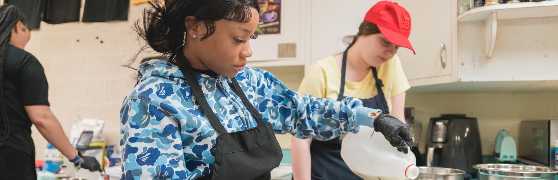 Person pouring milk into a measuring cup while another person observes in the background.