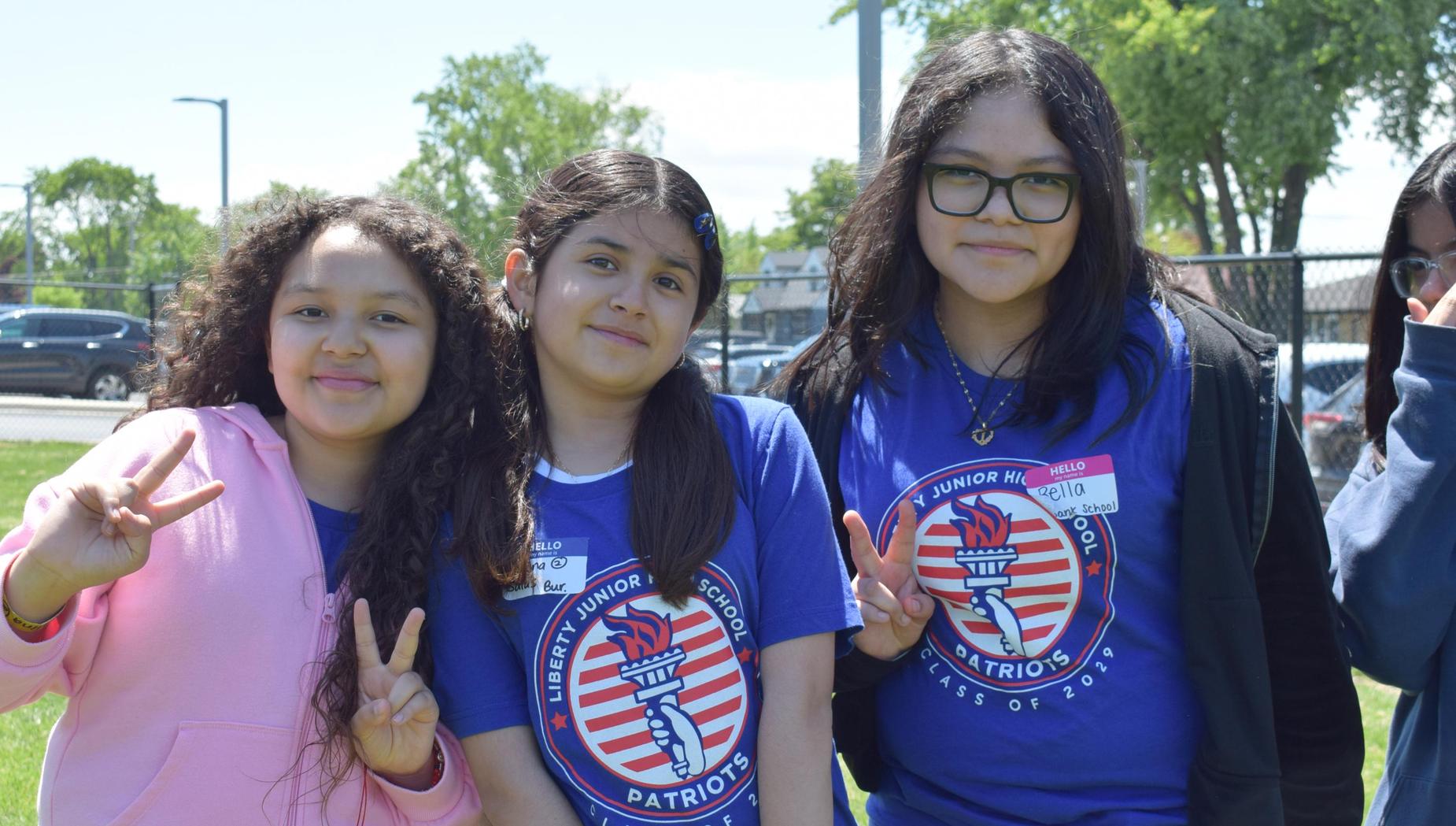 Three girls in blue shirts pose with peace signs on the grass.
