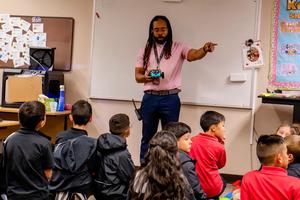 An instructor points while holding a game controller, addressing seated students.