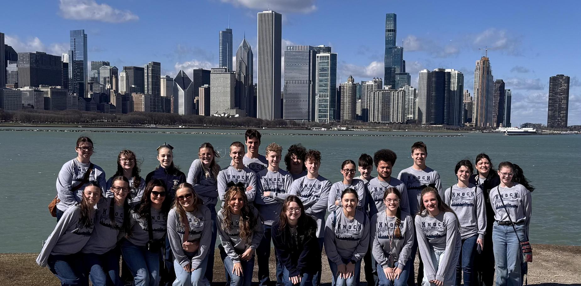 A group of students poses in front of a skyline with tall buildings by the waterfront.