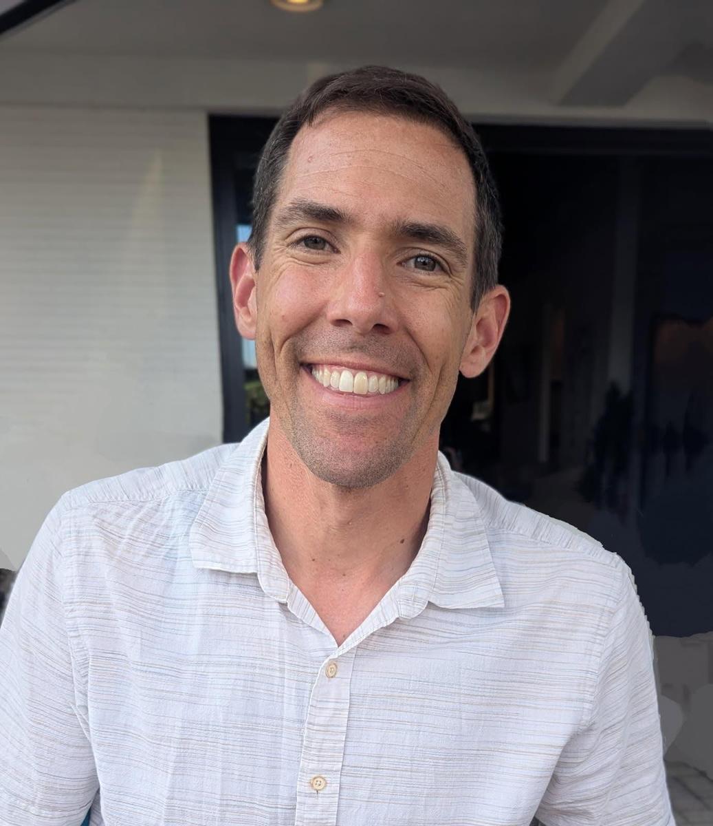 Smiling man in a light shirt poses outdoors, with a blurred background.