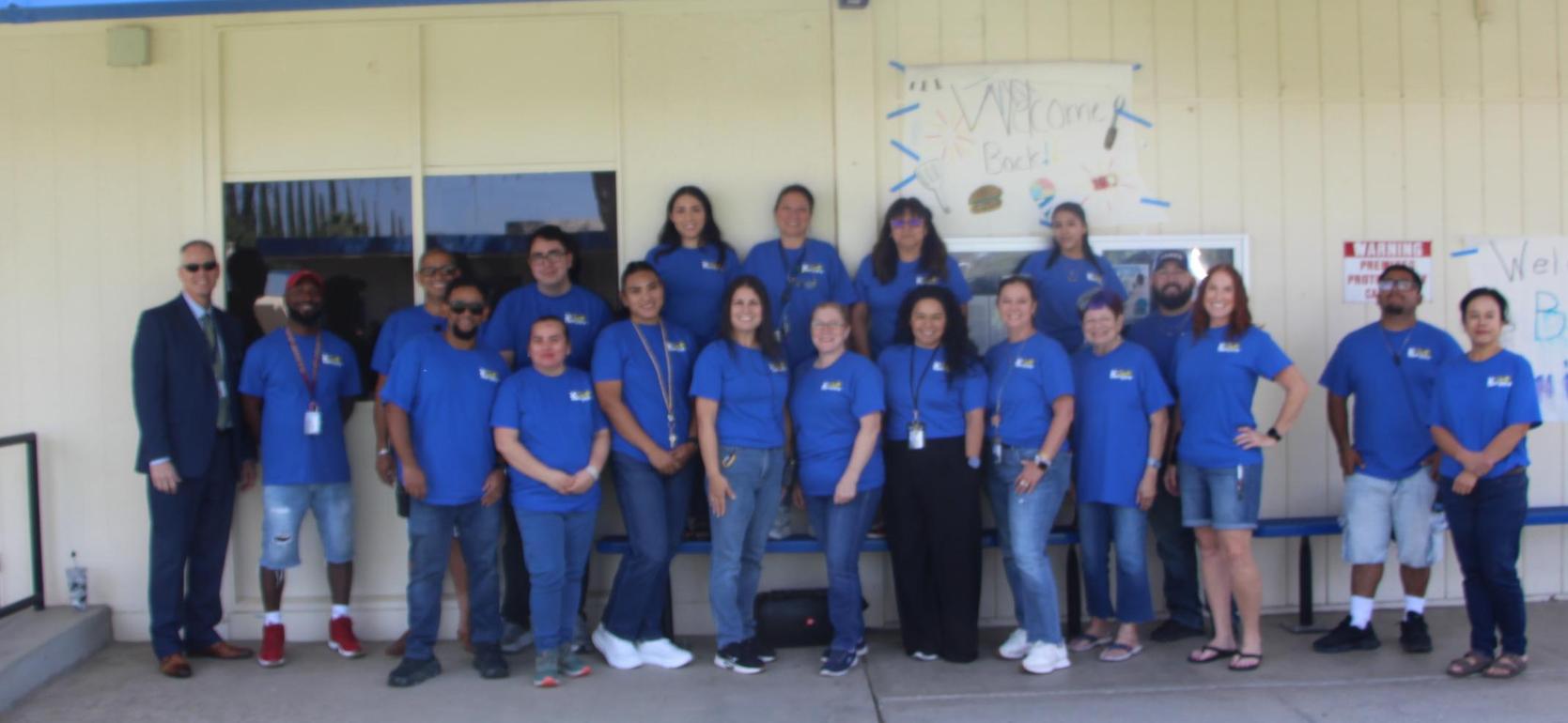 Group of people in blue shirts posing outside a school building with welcome signs.