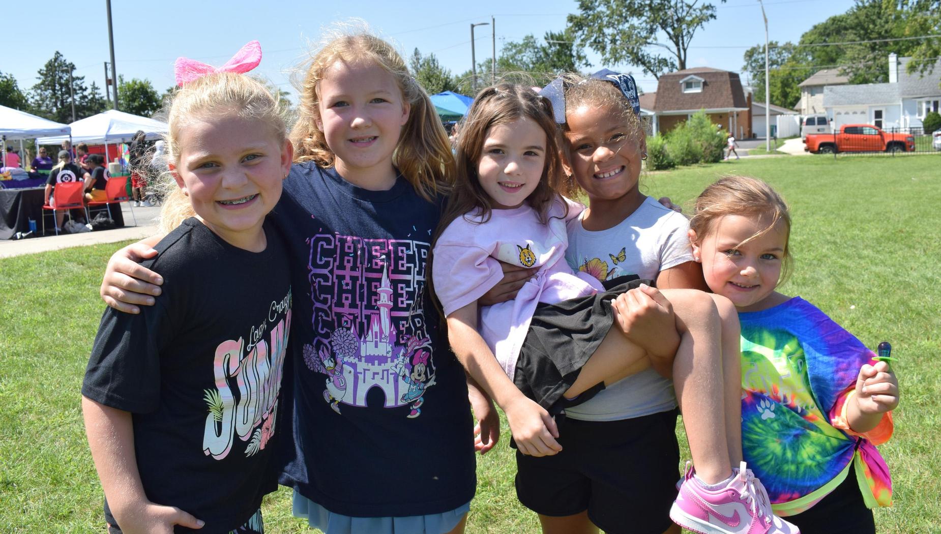 Five cheerful girls pose together outside, one being lifted off the ground.