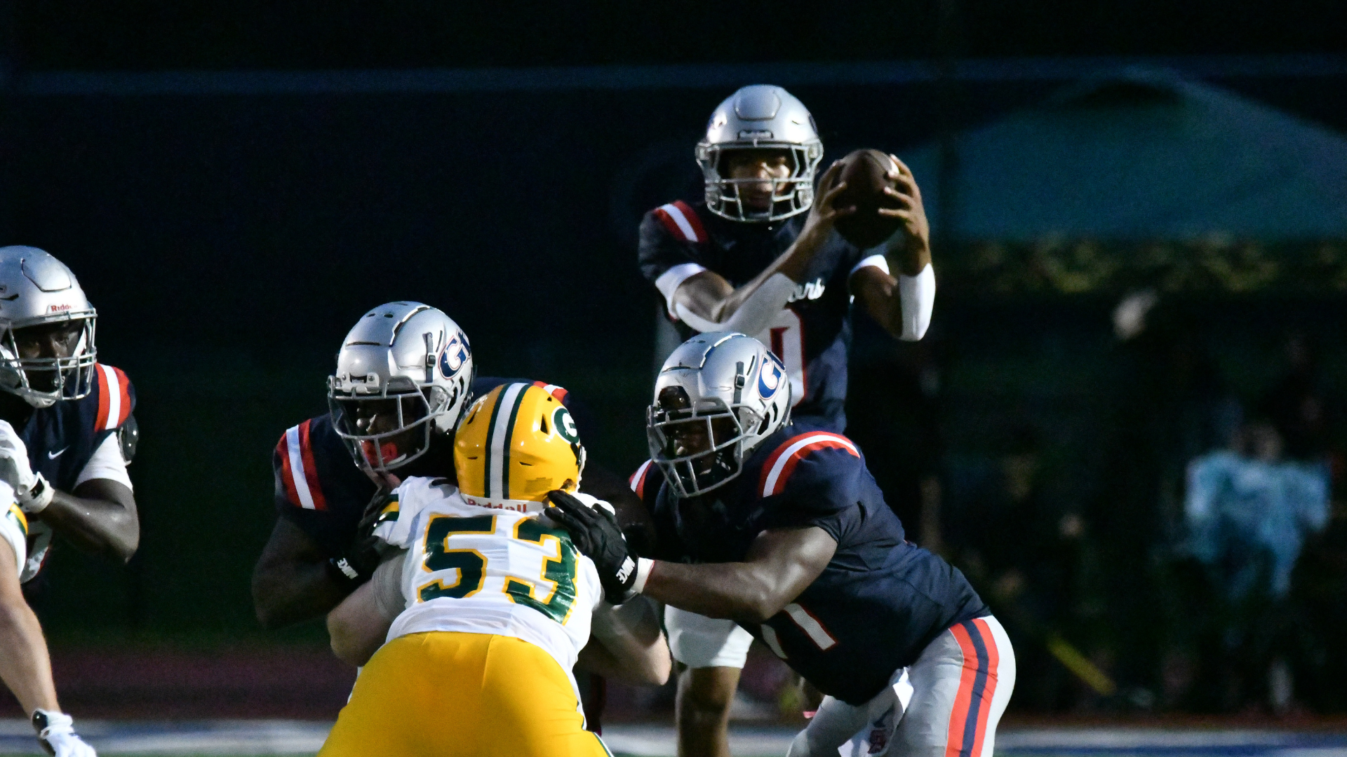 A football player in a dark uniform prepares to throw a pass while under pressure from defenders.