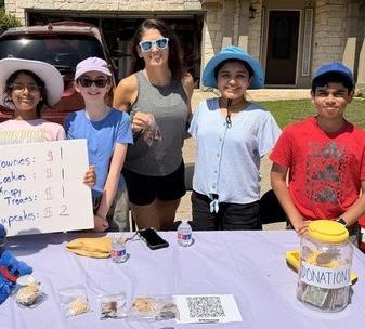 Students stand behind table with baked goods. Ms. Salazar stands with them.